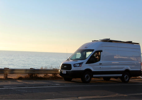 White Ford Transit camper van, an RV rental, parked on a coastal road overlooking the tranquil ocean at sunset.