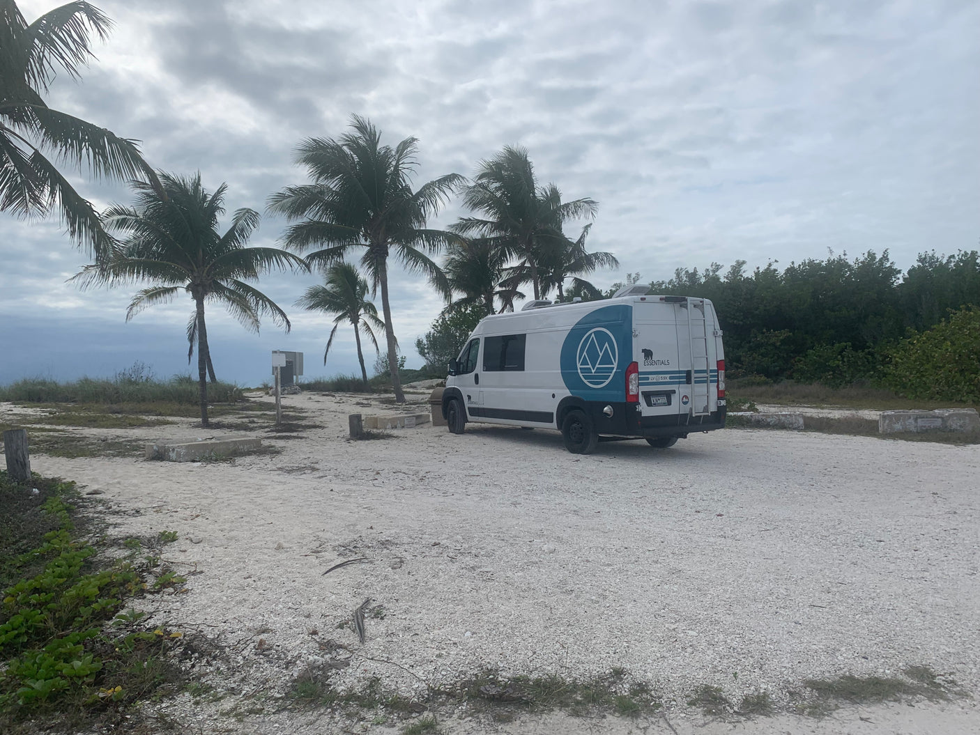 White **rv rental** van parked in a tropical setting. Palm trees and shell ground under cloudy skies, with the ocean in view.