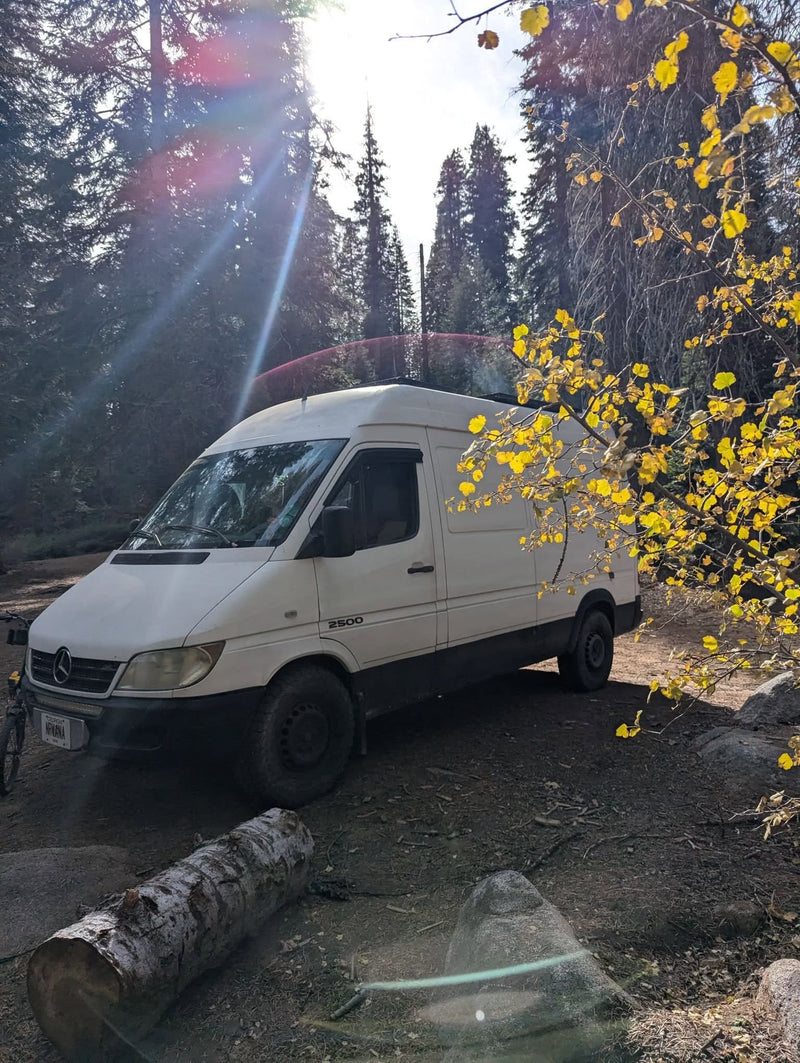 Experience van life! This white Mercedes Sprinter rv rental is perfectly parked in a sunny forest with bright yellow leaves.|Inside this RV rental camper van: a bed with blankets — photo 1
