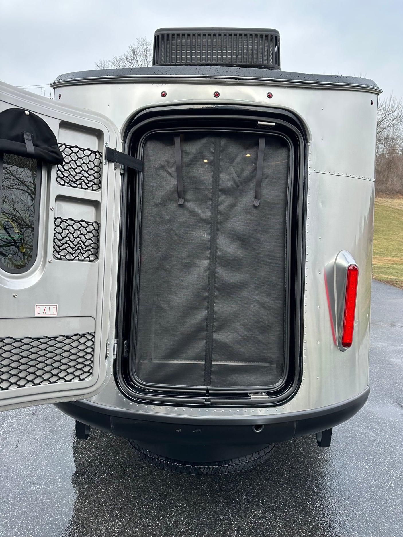 Rear view of an Airstream-style RV rental with its main door open, revealing a mesh screen, storage, & red taillight.