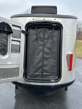 Rear view of an Airstream-style RV rental with its main door open, revealing a mesh screen, storage, & red taillight.
