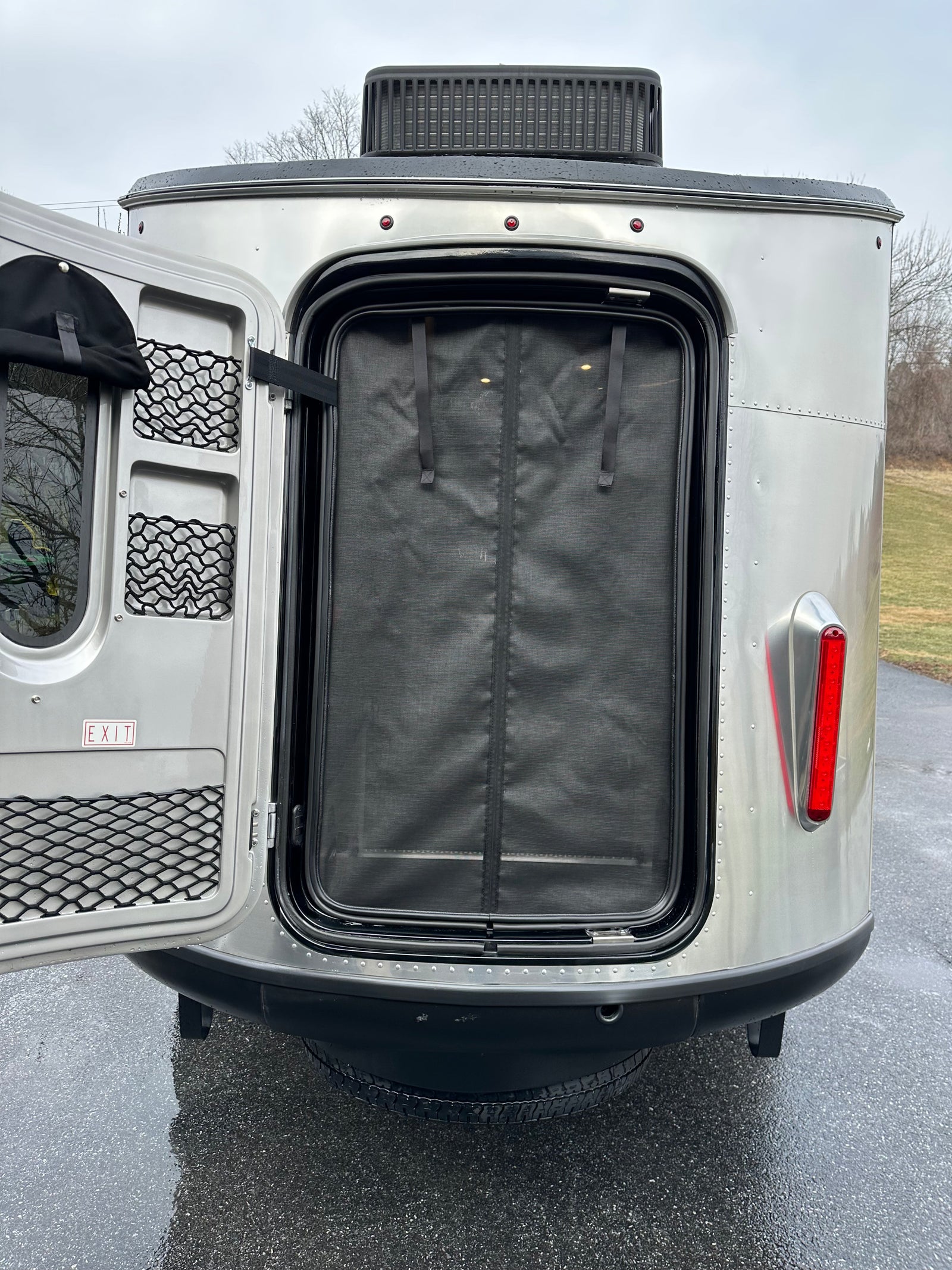 Rear view of an Airstream-style RV rental with its main door open, revealing a mesh screen, storage, & red taillight.