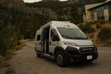 Silver rv rental camper van parked on a road shoulder with mountains, trees, and a house in the background.