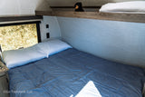 RV rental: Cozy bunk bed with white pillow & blue comforter. Window shows golden grass, upper bunk has a bear plush.