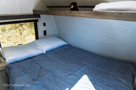 RV rental: Cozy bunk bed with white pillow & blue comforter. Window shows golden grass, upper bunk has a bear plush.