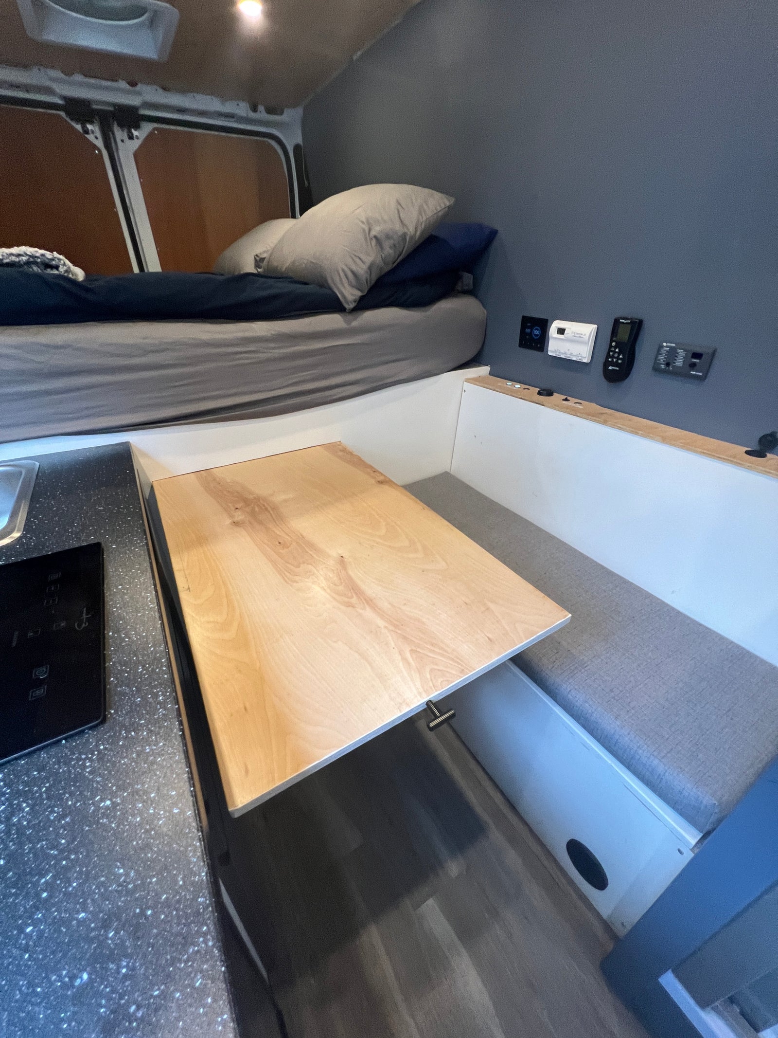 Compact RV rental interior featuring a bed, pull-out table with bench seating, and a small kitchen counter with a cooktop.