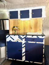 Modern RV rental interior: navy blue/gold geometric cabinets, wooden backsplash, black counter, and blue-doored overhead storage.