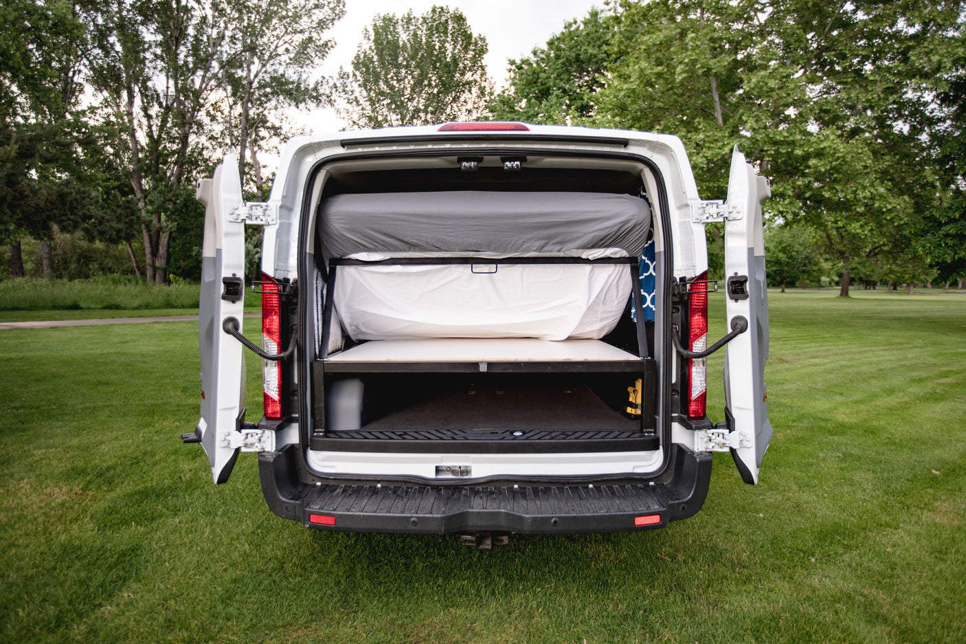 Explore with this RV rental! Rear of a white camper van with open doors, revealing a cozy two-level sleeping and gear storage area.
