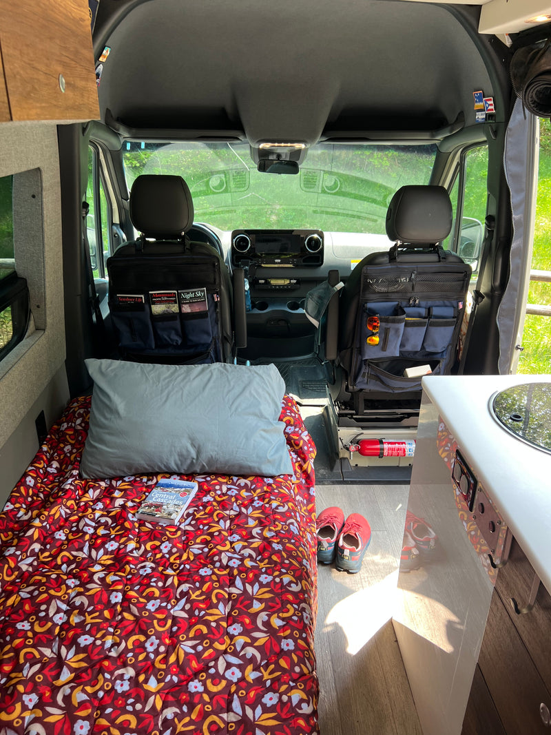 View into an rv rental showing a colorful bed, organized front seats, and the driver's cab leading to nature. — photo 17