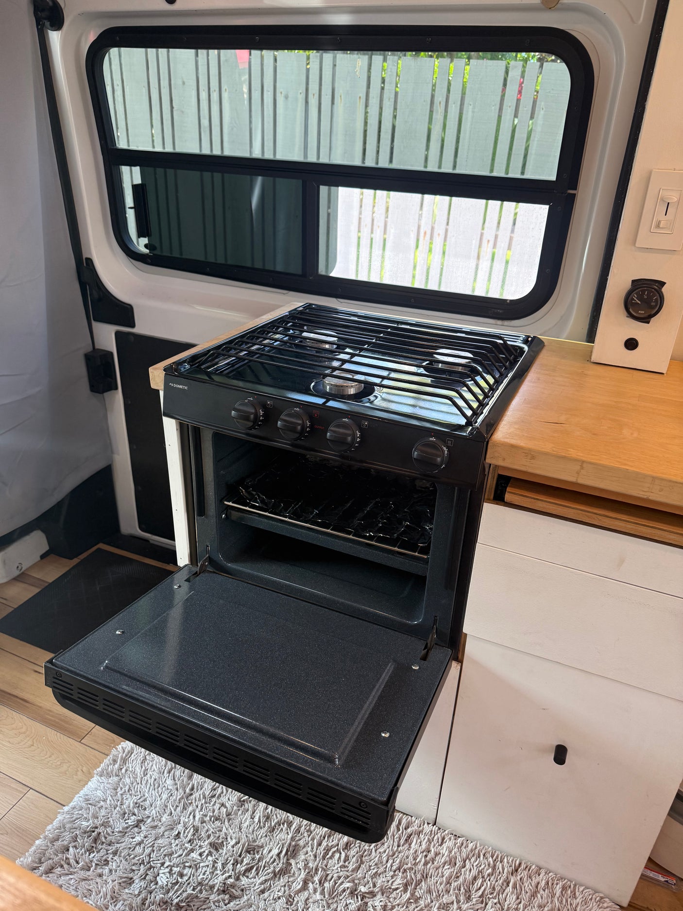 Inside an RV rental: a compact kitchen features a Dometic gas stove with oven, wooden counter, and window views.