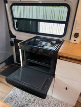 Inside an RV rental: a compact kitchen features a Dometic gas stove with oven, wooden counter, and window views.