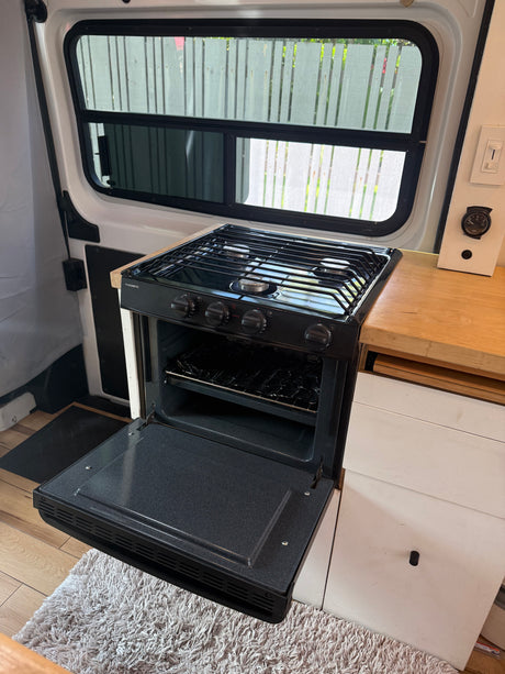 Inside an RV rental: a compact kitchen features a Dometic gas stove with oven, wooden counter, and window views.