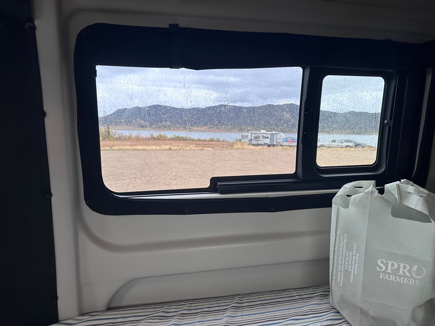 Rainy day inside our RV rental: Cozy view of a lake, distant mountains, and fellow campers through a droplet-covered window.