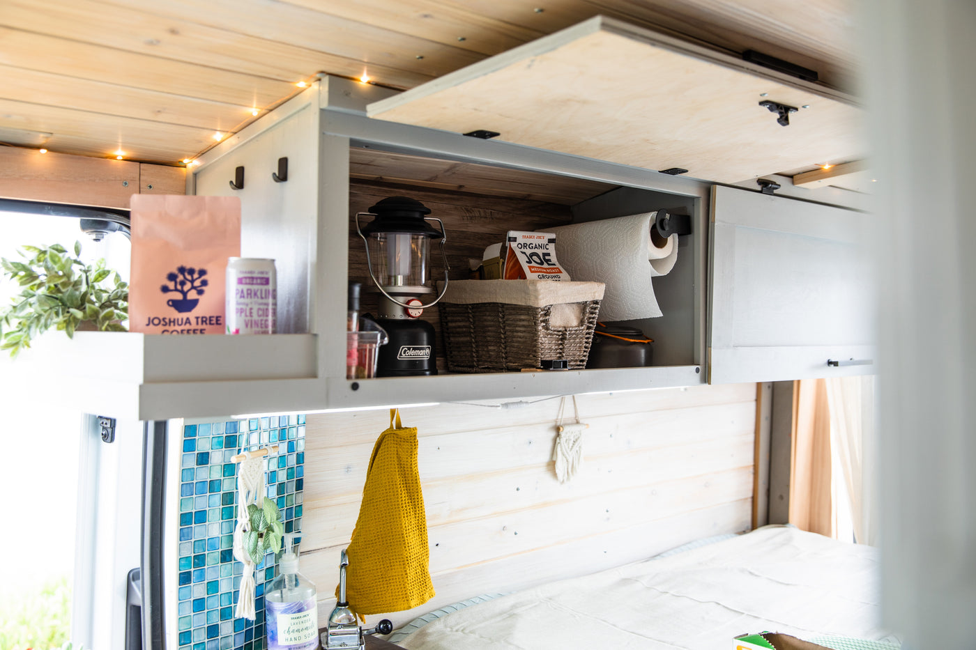 Cozy rv rental interior with wooden paneling & string lights. Overhead cabinets hold coffee, paper towels, and camping gear. — photo 15