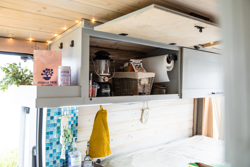 Cozy rv rental interior with wooden paneling & string lights. Overhead cabinets hold coffee, paper towels, and camping gear. — photo 15