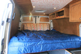 Inside an RV rental: Custom campervan interior featuring a bed with blue bedding, wood walls, cabinets, and a patterned ceiling.