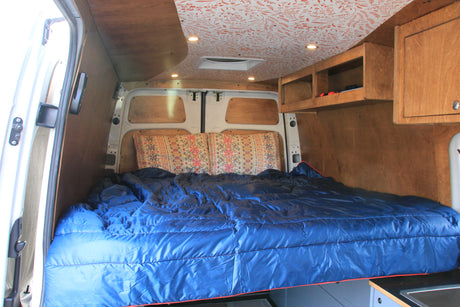 Inside an RV rental: Custom campervan interior featuring a bed with blue bedding, wood walls, cabinets, and a patterned ceiling.
