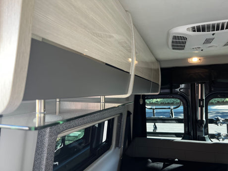 **RV rental** interior: light wood overhead cabinets, ceiling AC, and a clear glass shelf over a window with patterned trim.
