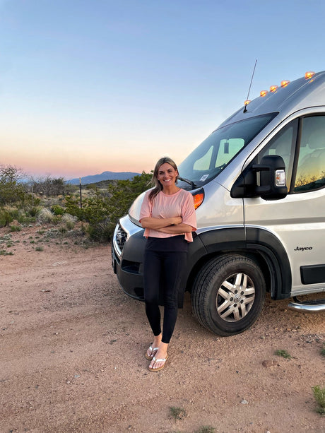 Smiling woman enjoys a beautiful desert sunset next to her Jayco RV rental camper van.