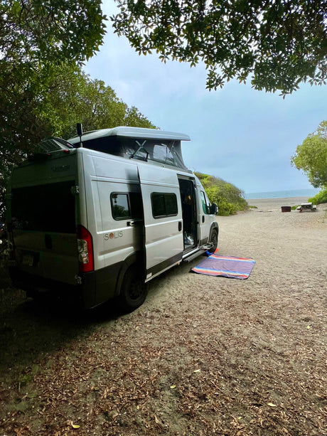 Coastal rv rental: A Solis campervan parked on sand, door open, mat out. Beach & trees for serene camping.