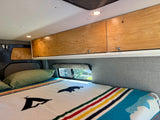 Cozy camper van RV rental with a comfy bed, illuminated wood cabinets, and a colorful striped blanket, ideal for travel.