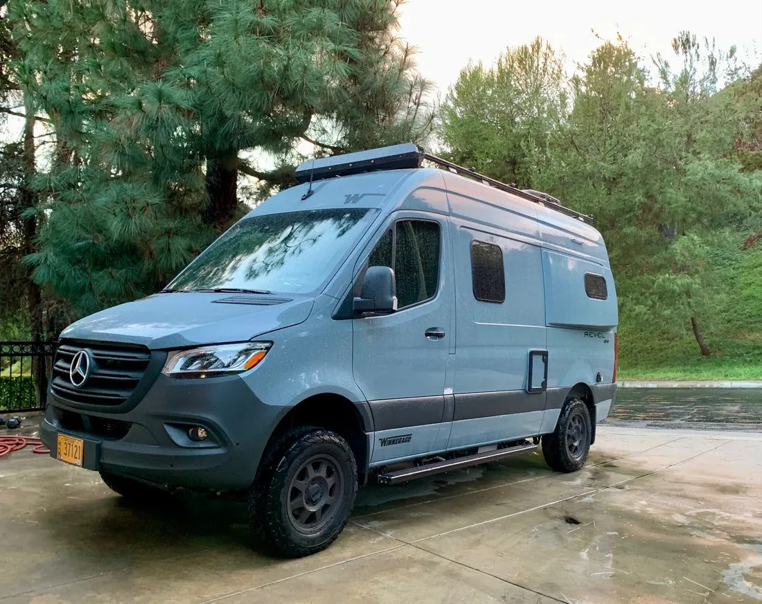 A grey Winnebago Revel 4x4 Mercedes Sprinter van, perfect for rv rental adventures, on a wet drive with trees.