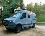 A grey Winnebago Revel 4x4 Mercedes Sprinter van, perfect for rv rental adventures, on a wet drive with trees.