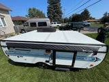 A white Jayco Jay Sport pop-up camper, with blue stripes & Dometic AC, sits on grass for RV rental.