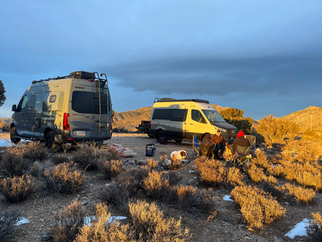 Two Winnebago Revel RV rental vans set up for boondocking. Campers and a dog gather around a warm campfire at sunset.