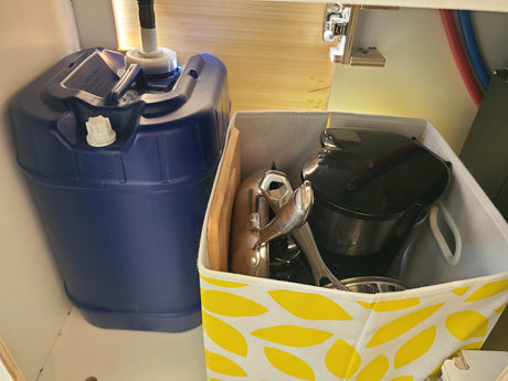 Inside an **RV rental**, a blue water jug with a pump sits next to a bin holding kitchen essentials for compact living.