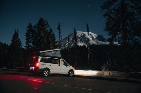RV rental camper van with pop-top parked at night. Starry sky above a snowy mountain and forest. Red taillights glow.