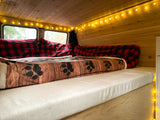 Cozy RV rental bedroom with wooden walls, string lights, and a bed featuring a paw print blanket and red plaid pillows.