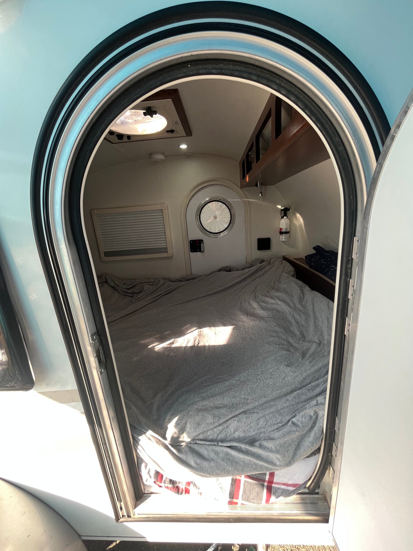 Step into a spacious RV rental bed area, seen through an arched door. Features include a comfy grey bed, small round door, and overhead storage.