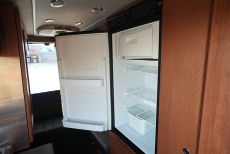 Open, empty refrigerator inside an RV rental, showing shelves, wood cabinetry, and a view to the outside.