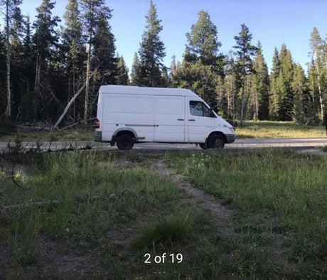 This white Sprinter van, ready for RV rental, is parked roadside in a scenic forest.