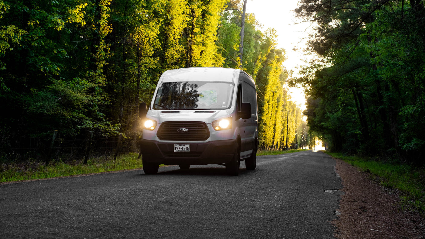 White Ford Transit van, ideal for RV rental, drives on a sun-dappled road through vibrant green trees. Explore nature!