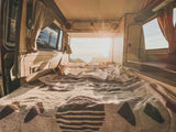 Cozy RV rental interior shows a made bed, patterned blankets, and a bright ocean sunset view from the rear.