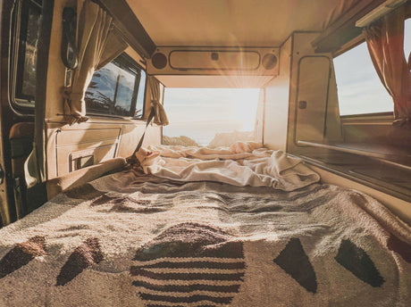 Cozy RV rental interior shows a made bed, patterned blankets, and a bright ocean sunset view from the rear.