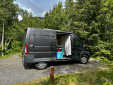 Dark gray rv rental van parked on gravel, side door open to reveal interior, surrounded by lush green trees and distant mountains.