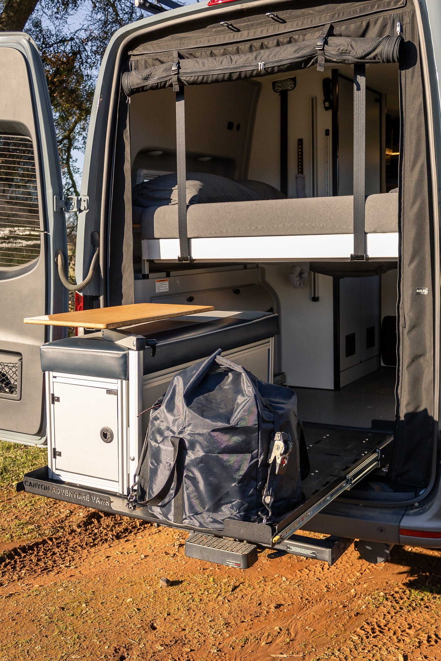RV rental camper van's open rear reveals a modular bed, side table, and pull-out storage with a duffel bag.