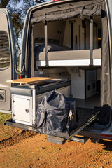 RV rental camper van's open rear reveals a modular bed, side table, and pull-out storage with a duffel bag.