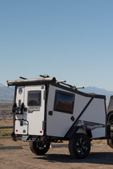A white and black TigerMoth overland trailer, ideal for an RV rental, parked on dirt with mountains in the background. Ready for off-road camping.
