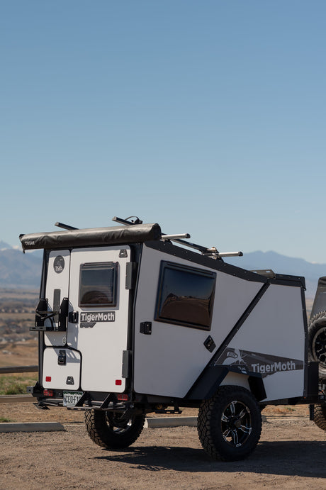 A white and black TigerMoth overland trailer, ideal for an RV rental, parked on dirt with mountains in the background. Ready for off-road camping.