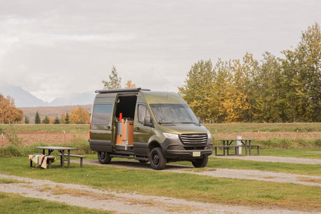 Green camper van RV rental parked at a scenic campsite with mountains & fall trees.