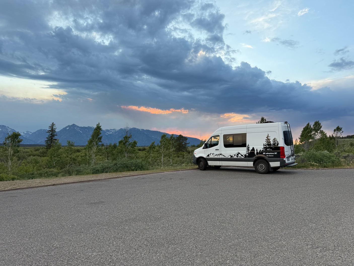 Scenic mountain views from this white RV rental van, parked roadside under a beautiful, cloudy sunset sky with orange hues.