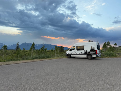 Scenic mountain views from this white RV rental van, parked roadside under a beautiful, cloudy sunset sky with orange hues.