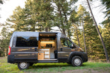 A grey camper van RV rental with its door open, showcasing a lighted custom kitchenette, parked amidst tall pine trees.