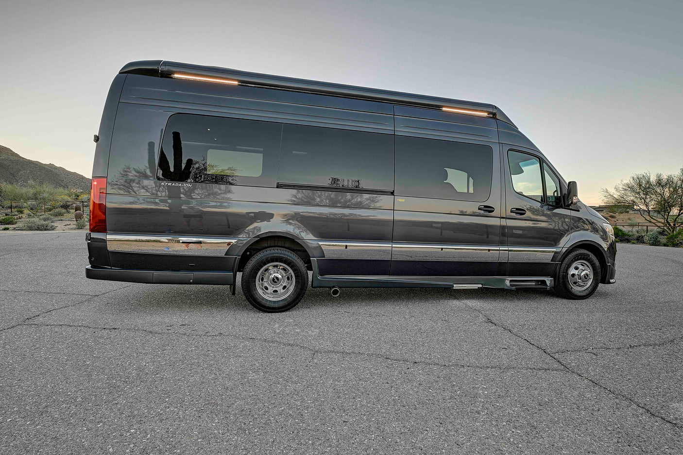 This sleek, dark grey Mercedes Sprinter camper van, ideal for an RV rental, is parked outdoors in a desert landscape with cacti reflections.