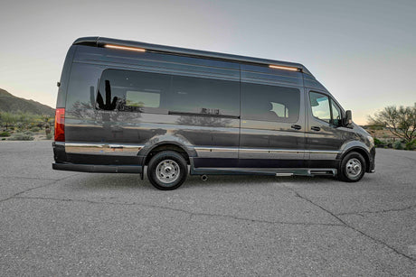 This sleek, dark grey Mercedes Sprinter camper van, ideal for an RV rental, is parked outdoors in a desert landscape with cacti reflections.