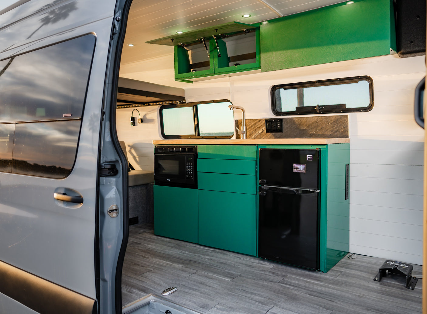 Inside an rv rental: modern kitchen with vibrant green cabinets, microwave, fridge, sink, and wood flooring visible from the open side door.